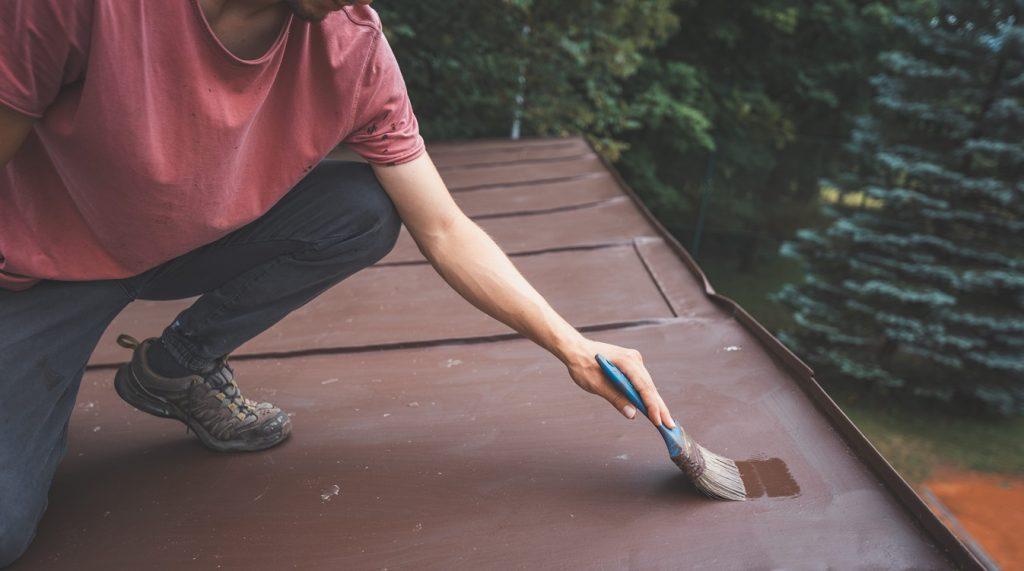 Man crouches down painting the surface of a roof with a small brush