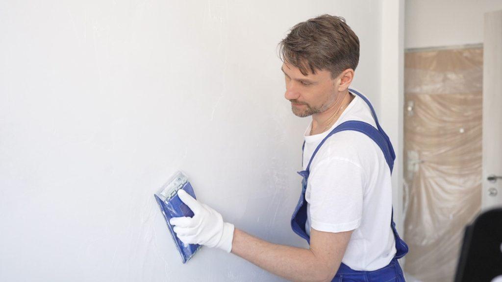 Construction worker wearing protective white gloves and blue construction coveralls is sanding