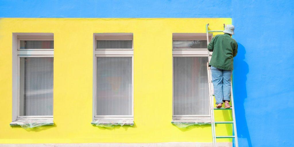 Female painter paints the wall of the facade of a building while standing on the stairs