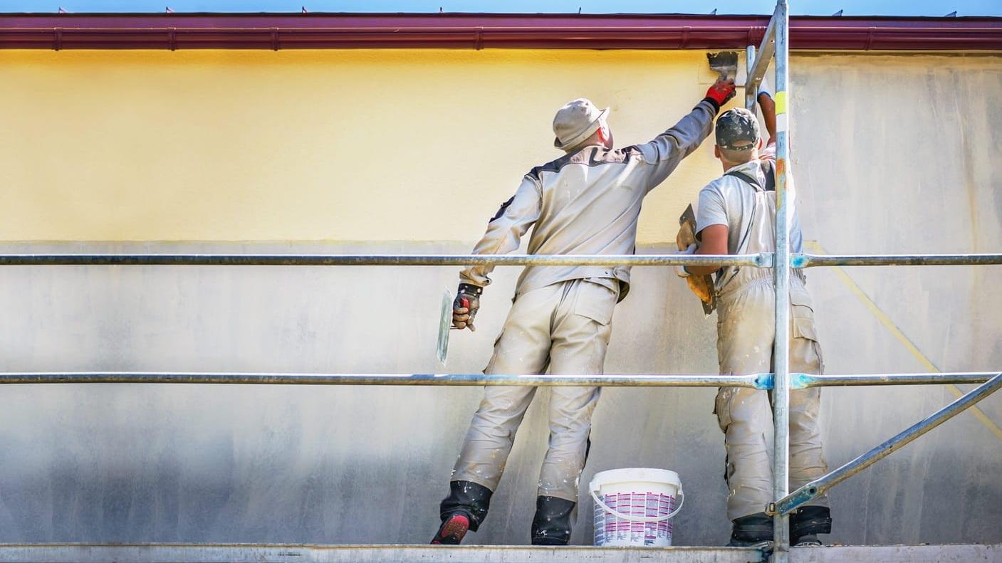 Two builders on safety scaffolding make yellow decorative plaster on the wall